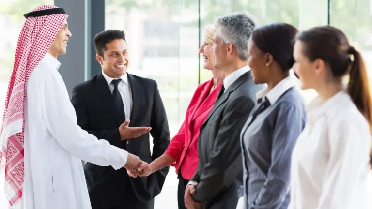 A group of business professionals from diverse backgrounds meeting and greeting in a modern office setting. A man in traditional Saudi attire shakes hands with a man in a suit, while other colleagues stand nearby smiling, indicating a successful business meeting or partnership.