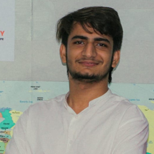 A young man with short brown hair and a trimmed beard, wearing a white shirt, standing in front of a world map with a slight smile.