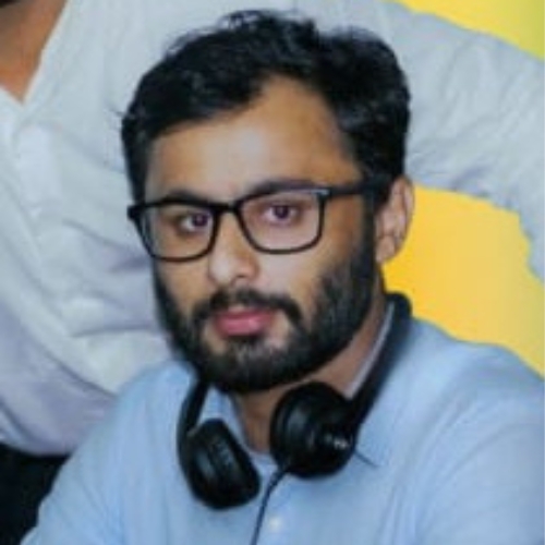 A man with short black hair, a full beard, and glasses, wearing a light blue shirt with black headphones around his neck.