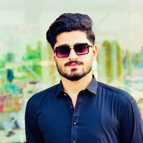 A man with styled black hair, a trimmed beard, and wearing dark sunglasses, dressed in a black button-up shirt, standing outdoors in bright sunlight